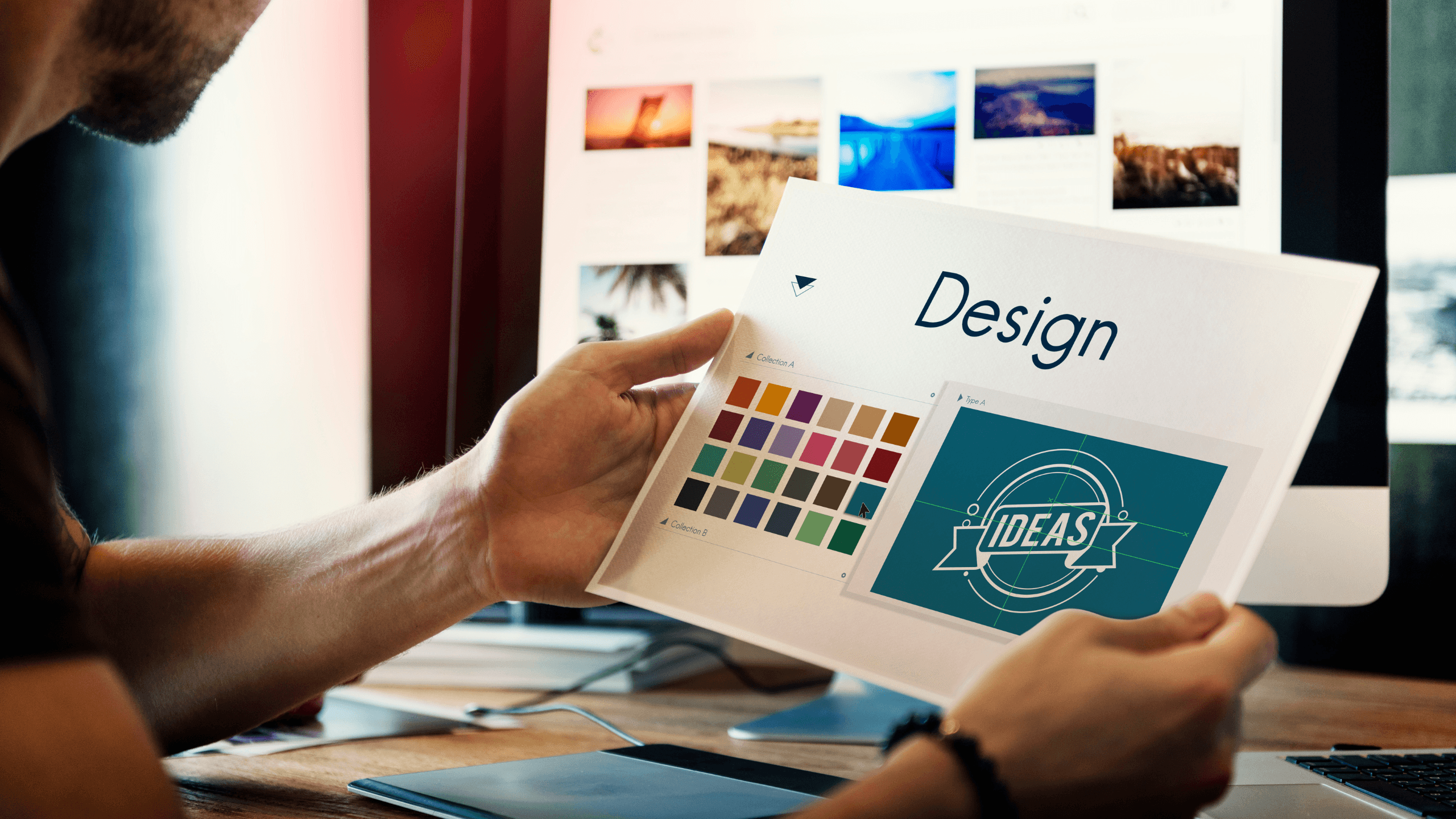A designer holding a printed design layout featuring color palettes and a logo concept, with a computer screen displaying visual design references in the background.